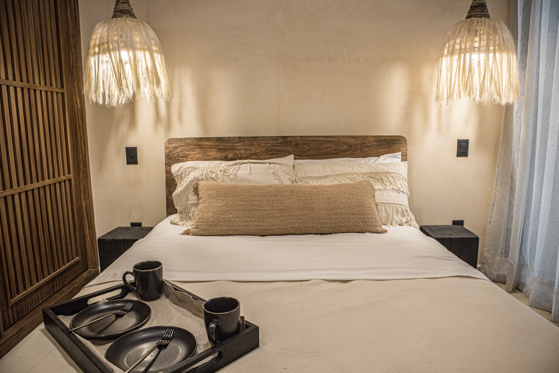 Vista frontal de Cama queen size de madera maciza con cojines decorativos bordados. Dos lámparas de techo de henequén y buró de madera