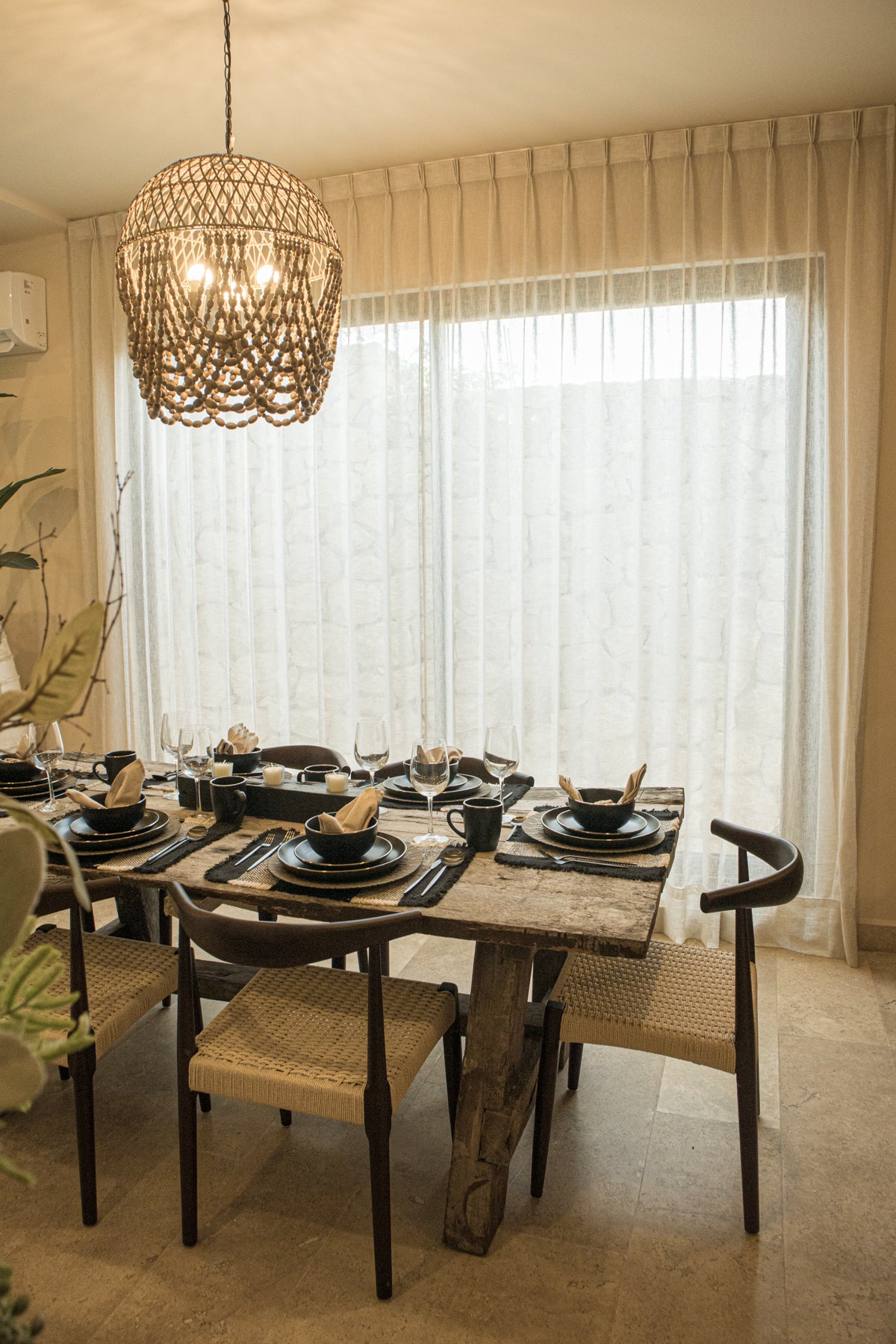 Comedor con mesa rústica de madera y sillas tejidas, decorado con vajilla negra y lámpara colgante. Encuentra muebles únicos en Prisma Muebles Tulum.