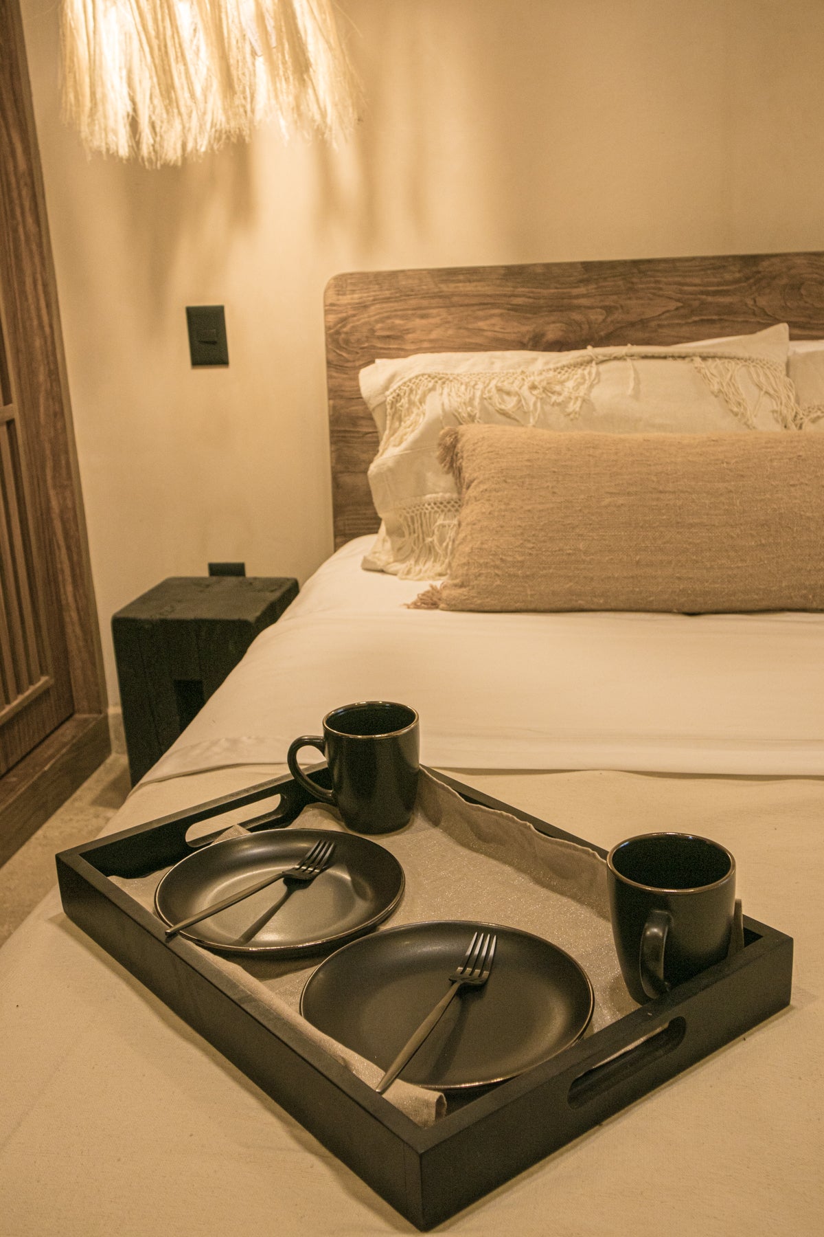 Habitación decorada con cabecera de madera, lámpara tejida y bandeja con vajilla negra sobre la cama. Encuentra muebles y decoración costera en Prisma Muebles Tulum.