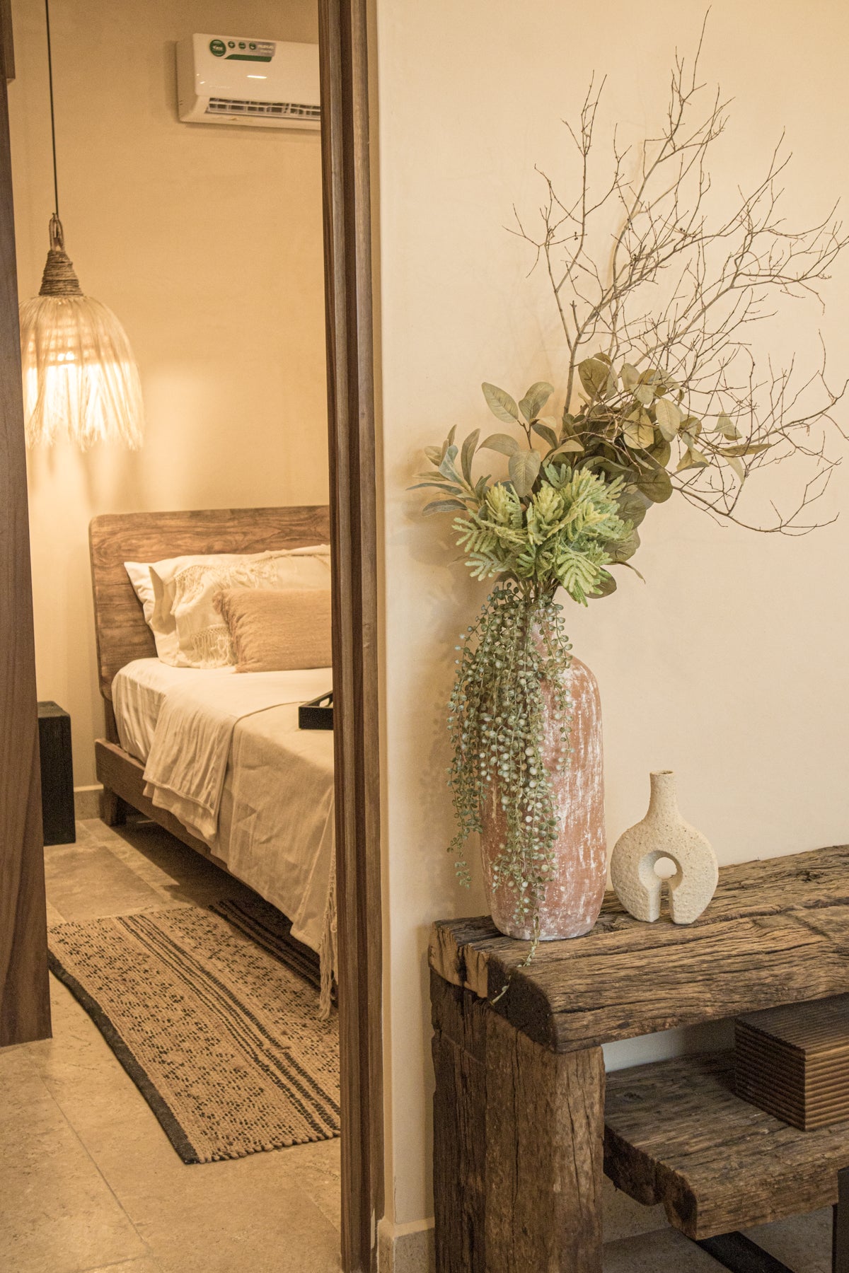 Entrada decorada con credenza vintage, jarrón artesanal con follaje y acceso a recámara con cama de madera y lámpara colgante. Prisma Muebles Tulum.