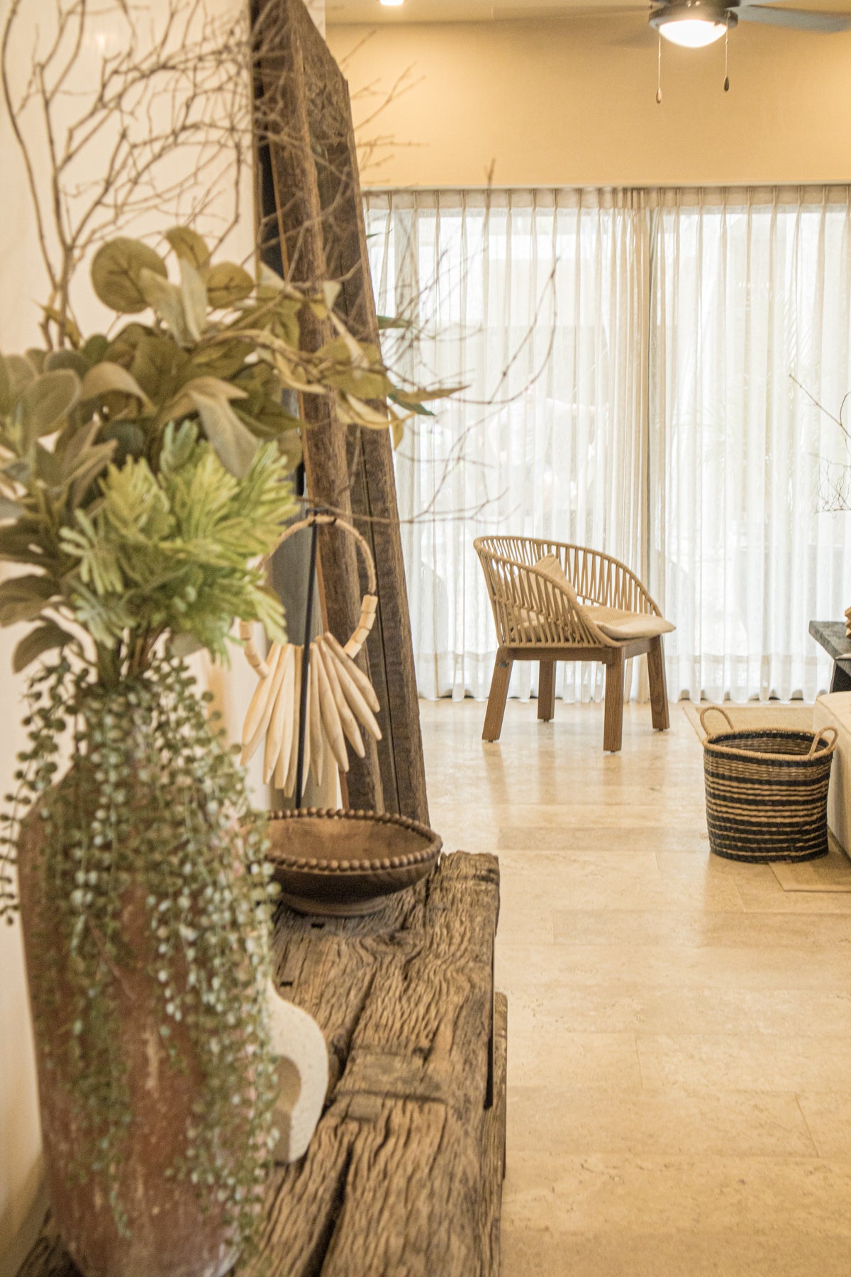 Decoración costera con mesa rústica de madera, jarrón con follaje verde, accesorios artesanales y silla de madera tejida al fondo. Encuentra esta decoración en Prisma Muebles Tulum.